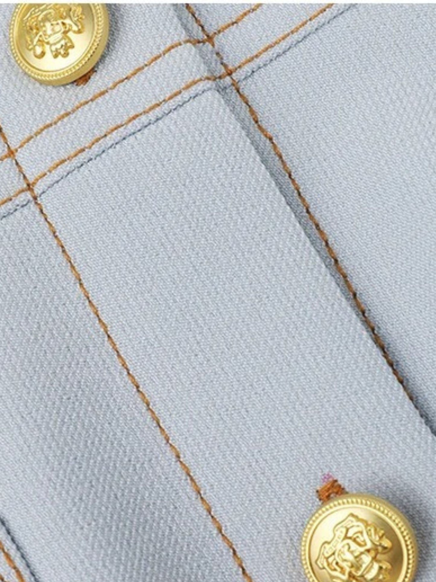 Detail of gold button and topstitching on light blue woven fabric.