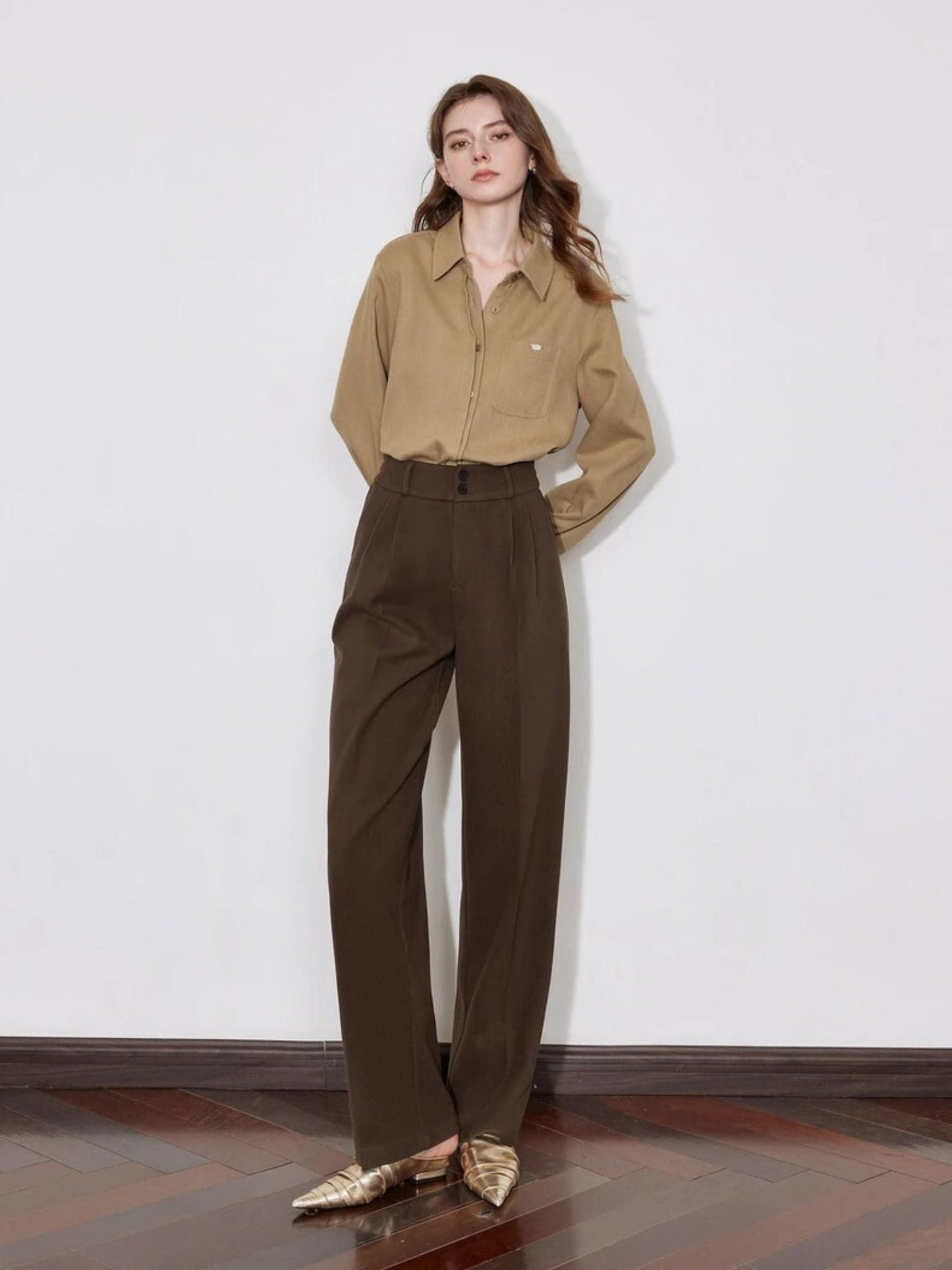 Model in brown pleated trousers with button-down shirt and gold shoes.