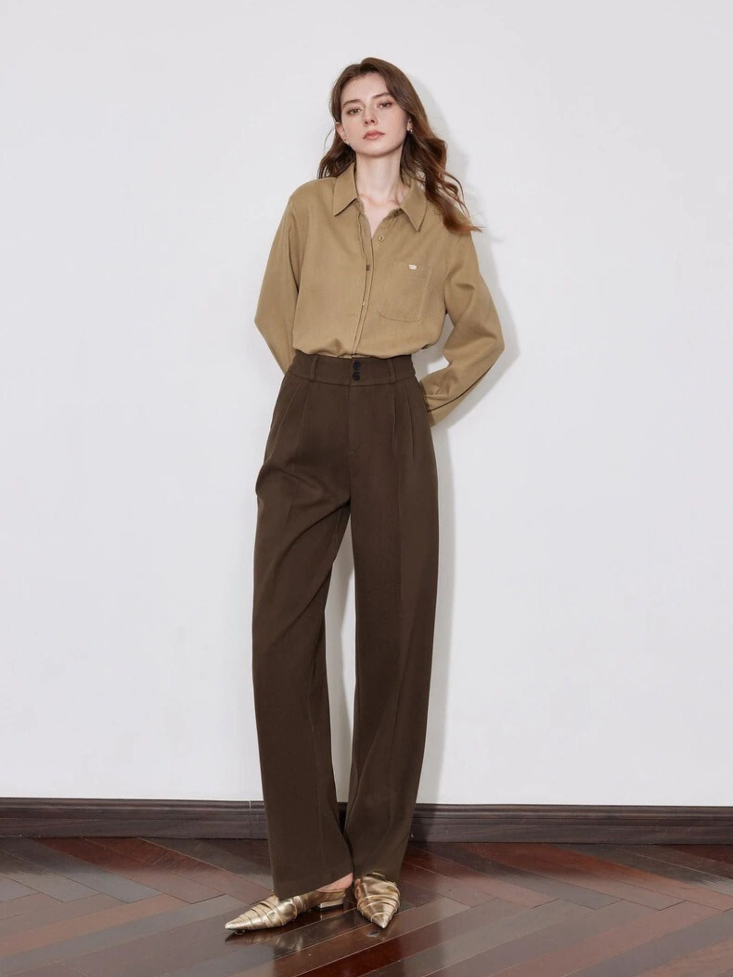 Model in brown pleated trousers with button-down shirt and gold shoes.