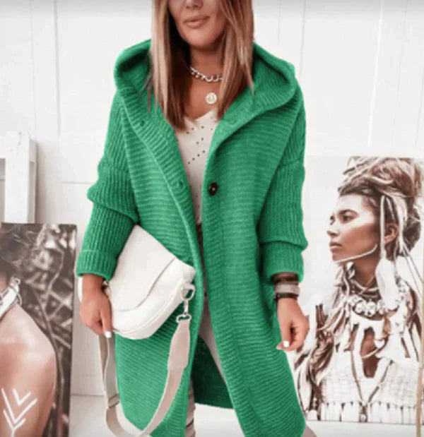 Eine Person trägt eine lange grüne Strickjacke mit Kapuze und zwei schwarzen Knöpfen und hält eine Tasche